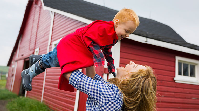 Mother lifting son in cape overhead outside barn