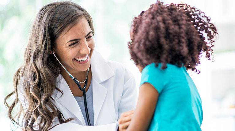 female doctor is indoors in a hospital room. She is wearing medical clothing and a stethoscope. She is checking the heart rate of a girl.