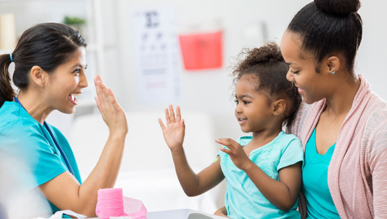 An African American mom holds her daughter as she high-fives a nurse.